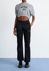 Felpa grigia corta con logo nero "NIKE 1972", abbinata a pantaloni neri con tasche laterali e piccolo swoosh Nike bianco, indossata con stivali neri.