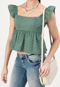 Top de algodón verde con escote cuadrado, mangas cortas con volantes y patrón de ojales; combinado con jeans azules desgastados y un bolso blanco.