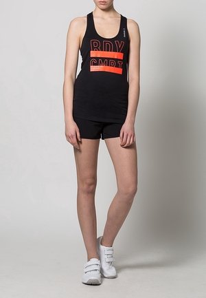 Canotta nera da allenamento con testo rosso "RDY CMPT", abbinata a pantaloni corti neri. Il modello indossa sneakers bianche con cinturini in Velcro. Texture liscia.