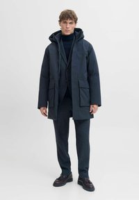 Tamsiai mėlynas parka su kapišonu, turintis dvi priekines kišenes, lygų paviršių ir tiesų siluetą, dėvimas virš tamsaus kostiumo ir golfuko.