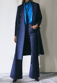 Long manteau bleu marine avec des boutons argentés, superposé sur un haut bleu vif et un jean évasé noir. Tissu lisse et design structuré avec une coupe ajustée.