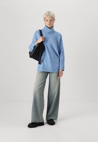 Un pull col roulé tricoté bleu clair, coupe ample, associé à un jean en denim délavé à jambes larges et des chaussures à plateforme noires, portant un sac fourre-tout noir.