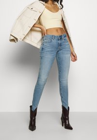 Mujer con jeans skinny de color azul claro, botas de tacón negras, top corto beige y chaqueta de mezclilla crema sobre los hombros.