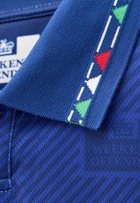 Col de vêtement bleu avec motifs triangulaires brodés en vert, rouge et blanc, et étiquette de marque partiellement visible sur un tissu texturé.