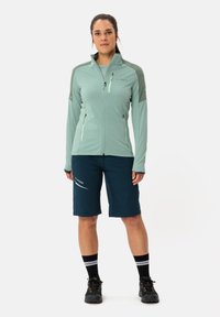 Lichtmoosfarbene Zip-Jacke mit dunkelgrünen Akzenten, kombiniert mit navyblauen Shorts. Besitzt robusten Stoff, Taschen und kontrastierende Streifen an den Socken.