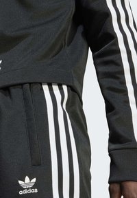 Fato de treino desportivo preto com detalhes em branco nas mangas e pernas, feito de tecido texturizado e com um logo da Adidas no peito.