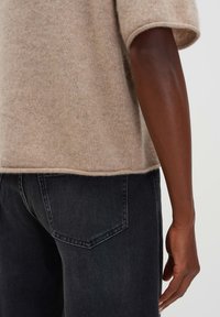 Persona di pelle scura che indossa un maglione beige a maniche corte e jeans in denim neri, con un'inquadratura ravvicinata sulla zona del braccio e della tasca posteriore.