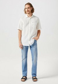 Koszula z krótkim rękawem, biała, zapinana na guziki, z kieszenią, w połączeniu z jasnoniebieskimi dżinsami i czarnymi sandałami. Gładki materiał, luźny fason.