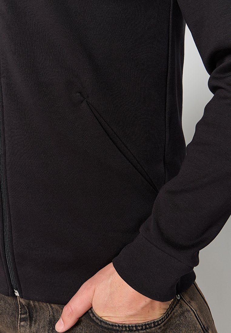 Ingrandimento di una persona che indossa una giacca nera con zip e una tasca diagonale, con la mano che riposa nella tasca dei pantaloni in denim marroni.