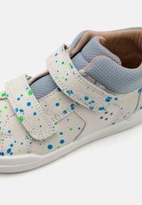 Sneaker mit einem weißen Lederäußeren, das mit blauen und grünen Spritzmustern versehen ist. Klettverschlussriemen und hellgraue Stoffakzente am Kragen.
