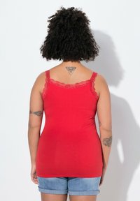 Top rosso senza maniche con bordi in pizzo, design aderente, tessuto morbido e spalline sottili. Il modello indossa pantaloncini di jeans; tatuaggi visibili su braccia e schiena.