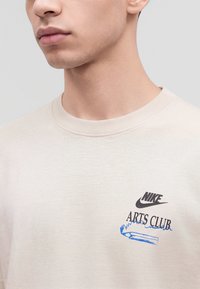 Мъж, носещ светлобежова тениска на Nike с текст "Arts Club" и синя графика на молив в лявата част на гърдите.