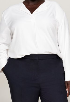 Personne portant un chemisier blanc ample à manches longues, rentré dans un pantalon bleu marine foncé taille haute, une main dans la poche.