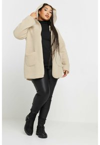 Beige fleecejacka med huva, två framfickor och avslappnad passform, ihop med svarta läderleggings och robusta svarta stövlar.