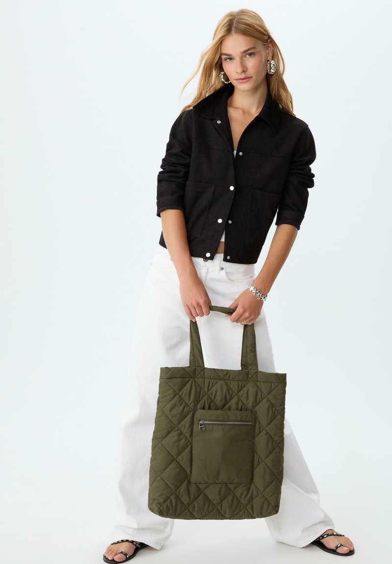 Gestepptes olivgrünes Tote-Bag mit einer vorderen Reißverschlusstasche und robusten Trägern; kombiniert mit einer schwarzen Wildlederjacke und weißen, weiten Hosen.