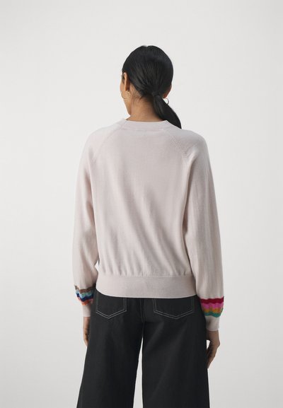 Pull léger rose avec des poignets côtelés, présentant des rayures multicolores, coupe décontractée et texture douce ; porté par-dessus un pantalon large sombre.