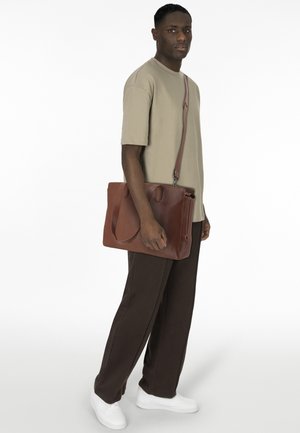 Sac en bandoulière en cuir brun avec une texture lisse et des poignées supérieures, porté en travers du corps, associé à une chemise beige et un pantalon sombre.