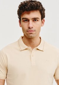 Beżowa koszulka polo o miękkiej fakturze, z krótkimi rękawami i klasycznym kołnierzykiem. Zawiera subtelną naszywkę z logo oraz dwukluczowy zamek.