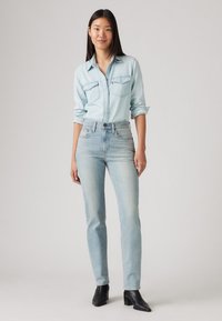 Šviesiai mėlyna džinsinė marškinė su dviem priekinių kišenėmis, derinamos su šviesiai plovtais džinsais. Modelis dėvi juodus kulkšninius batus su stambiu kulnu.