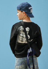 Garçon portant une casquette bleue à l'envers, un sweat-shirt noir avec un motif d'ours brillant, un jean en denim clair, et un sac bandoulière noir avec un bandana bleu.