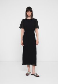 Svart midi-kjole med korte, gjennomsiktige ermer, teksturert stoff, tettsittende midje og rett kant. Kombinert med svarte og hvite mønstrede sandaler.