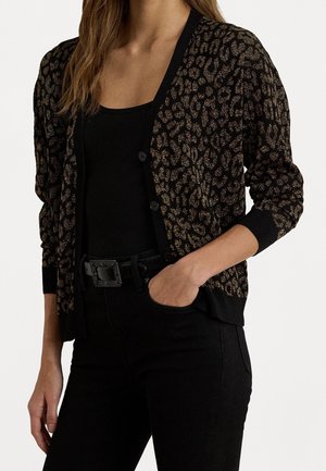 Femme portant un haut noir et un pantalon noir taille haute avec un cardigan texturé noir et or imprimé léopard, une main dans la poche.