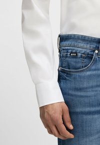 White long-sleeve shirt cuff contrasted against blue denim jeans. Jeans feature a black label with "BOSS" and visible stitching details.