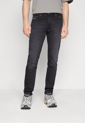 Homme portant un jean slim noir et des chaussures de sport grises debout devant un fond clair uni.