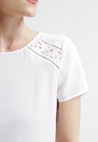 Camicia bianca con maniche corte, caratterizzata da un pannello in pizzo sulla spalla e cuciture decorative, texture liscia e scollatura rotonda.