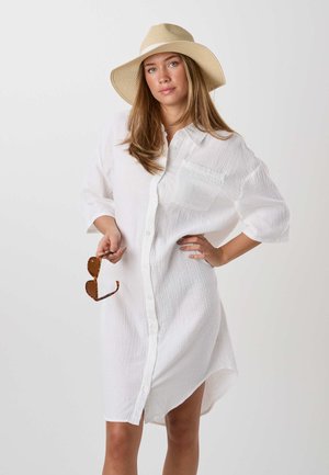 Kvinde iført en hvid knap-op kjole og en beige hat med bred skygge, der holder skildpaddeskal-solbriller med den ene hånd på hoften.