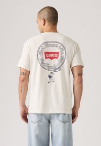 T-shirt à manches courtes couleur crème avec un motif graphique bleu marine au dos, comprenant le logo Levi's et un motif de corde. Coupe décontractée.
