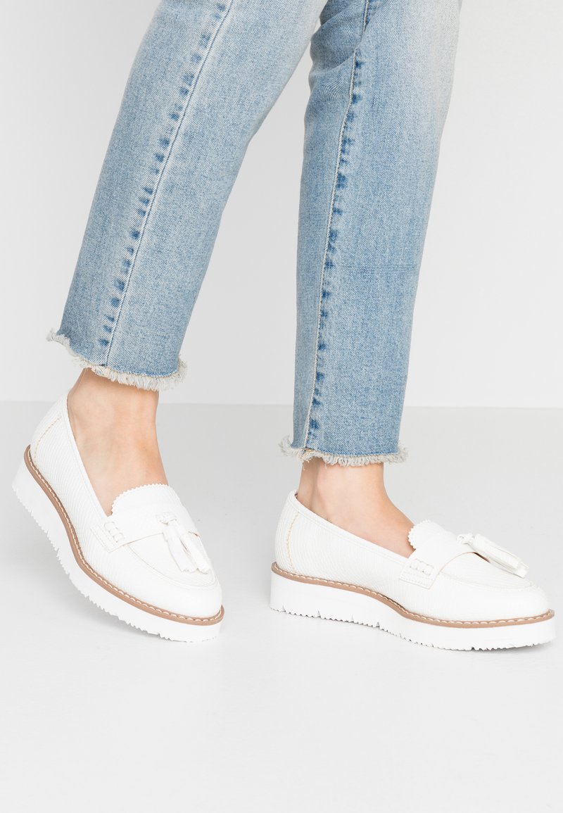 Hvite loafers med teksturert overdel, med en duskdetalj og en kontrasterende brun såle. Brukt sammen med lyseblå jeans med frynser.