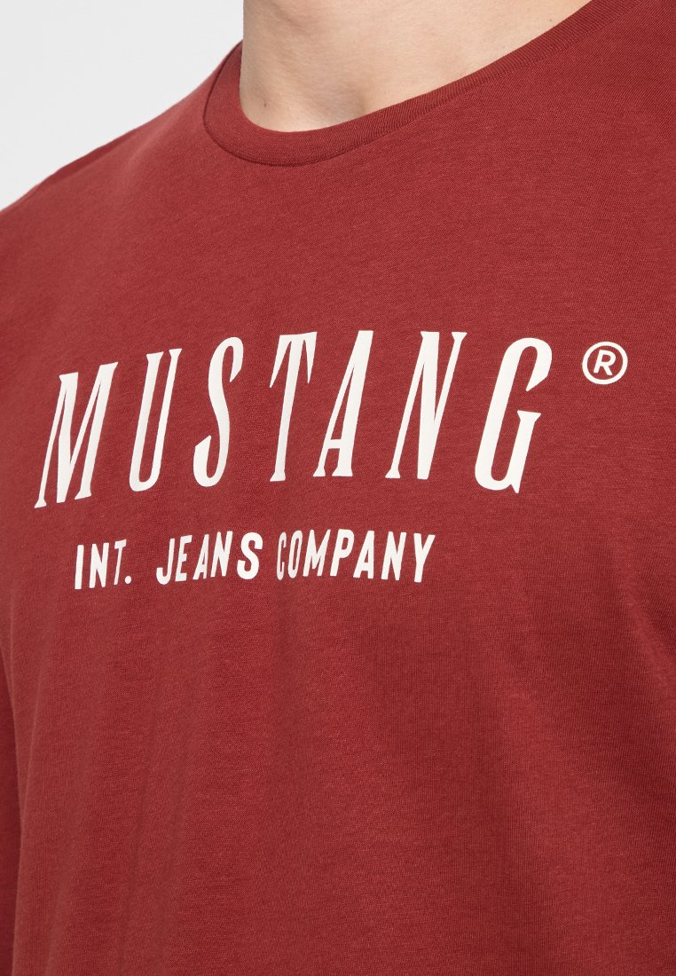 Czerwona bawełniana koszulka z okrągłym dekoltem, krótkimi rękawami i wyraźnym białym napisem "MUSTANG INT. JEANS COMPANY." Gładka tekstura.