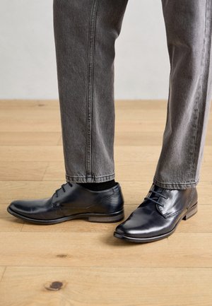 Zapatos de vestir de cuero negro usados con jeans grises de pie sobre suelo de madera.