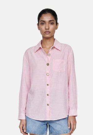Donna con capelli scuri indossa una camicia rosa chiaro a maniche lunghe con bottoni e pantaloncini di jeans azzurri su uno sfondo bianco uniforme.