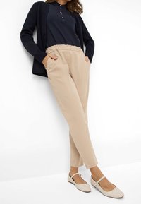 Pantalon beige à taille élastique avec poches latérales, associé à un polo bleu marine et un cardigan noir. Chaussures plates de couleur claire complètent la tenue.