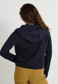 Personne avec de longs cheveux bruns portant un sweat à capuche matelassé bleu marine et un pantalon jaune moutarde, vu de dos contre un fond uni.