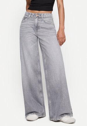 Mujer con vaqueros gris claro de talle alto y pierna ancha, combinados con zapatillas blancas y una camiseta corta negra, de pie frente a un fondo liso.