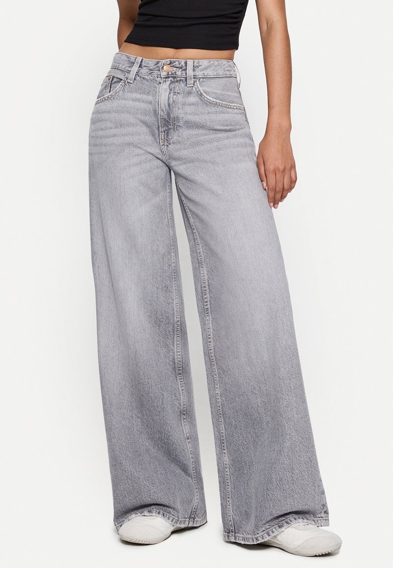 Femme portant un jean gris clair taille haute à jambes larges, associé à des baskets blanches et un top noir court, debout devant un fond uni.