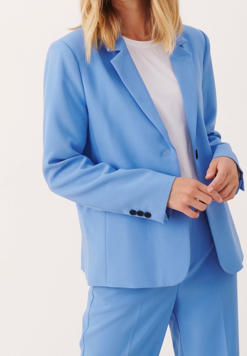 Femme portant un blazer bleu clair et un pantalon assorti par-dessus une chemise blanche, ajustant les manches du blazer devant un fond blanc.