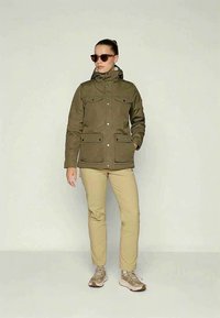 Kvinde iført en olivengrøn hættejakke, beige bukser, brune sneakers og solbriller, stående med den ene hånd i lommen mod en ensfarvet baggrund.