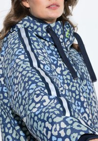 Voděodolná bunda s modro-zeleným abstraktním vzorem, kapucí a černými šňůrkami, s kontrastními bílými pruhy na rukávech.