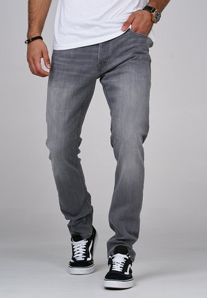 Homme portant un jean gris, des baskets blanches avec des touches noires, un t-shirt blanc, une montre-bracelet et un bracelet, une main dans la poche.