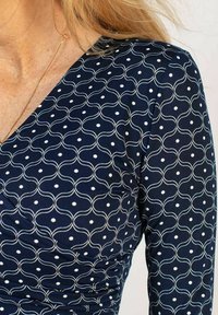 Marinblus med en vågig vit mönstring och små prickar. Designen har en figurnära silhuett och subtil textur med samlingar på sidan.