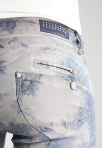 Vaqueros de mezclilla azul claro con un patrón floral, bolsillo con cremallera en la parte posterior y costuras decorativas. Parche de cuero con la marca en la cintura.