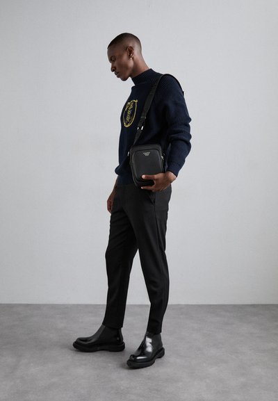 Svart crossbody-väska med rektangulär form och slät yta, bärs med en marinblå tröja med en gul emblem och svarta slim-fit-byxor.