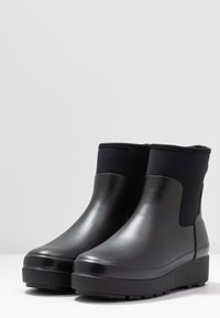 Une paire de bottes de cheville noires imperméables avec une finition mate lisse et des semelles en caoutchouc épaisses sur une surface blanche et un fond gris.
