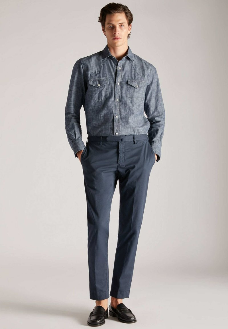 Man die met zijn handen in zijn zakken staat, gekleed in een blauwe chambray shirt, marineblauwe broek en zwarte instapschoenen tegen een effen achtergrond.