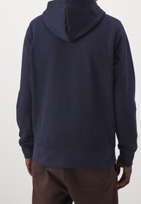 Hombre vistiendo una sudadera con capucha azul marino lisa y pantalones marrones, visto desde atrás contra un fondo claro.