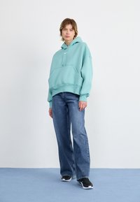 Moletom oversized verde menta com cordões, bolso frontal; combinado com calças jeans azuis largas e ténis pretos, em um fundo neutro.